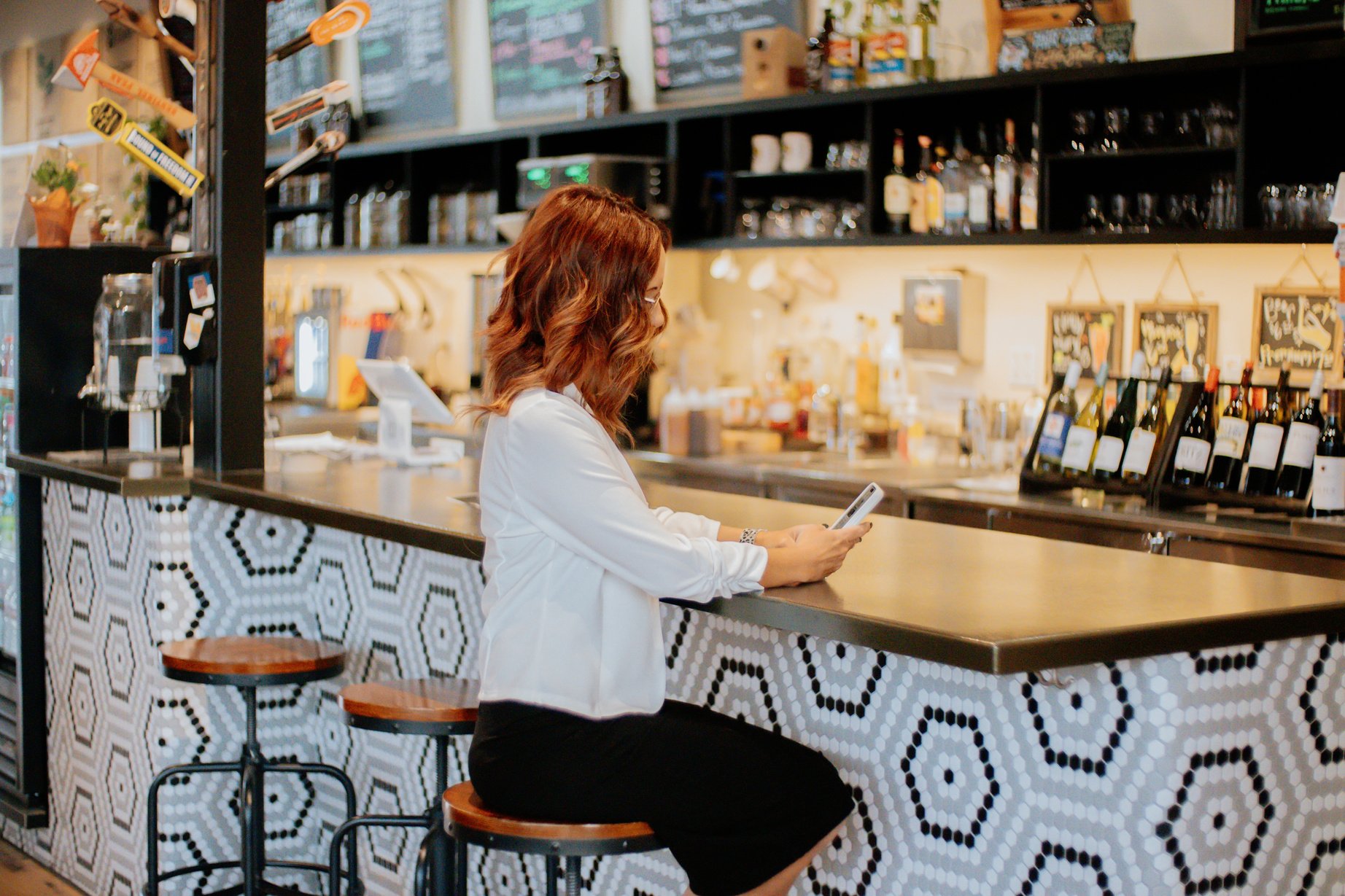 A woman in a white blazer and black skirt sitting at a bar and looking at her phone.